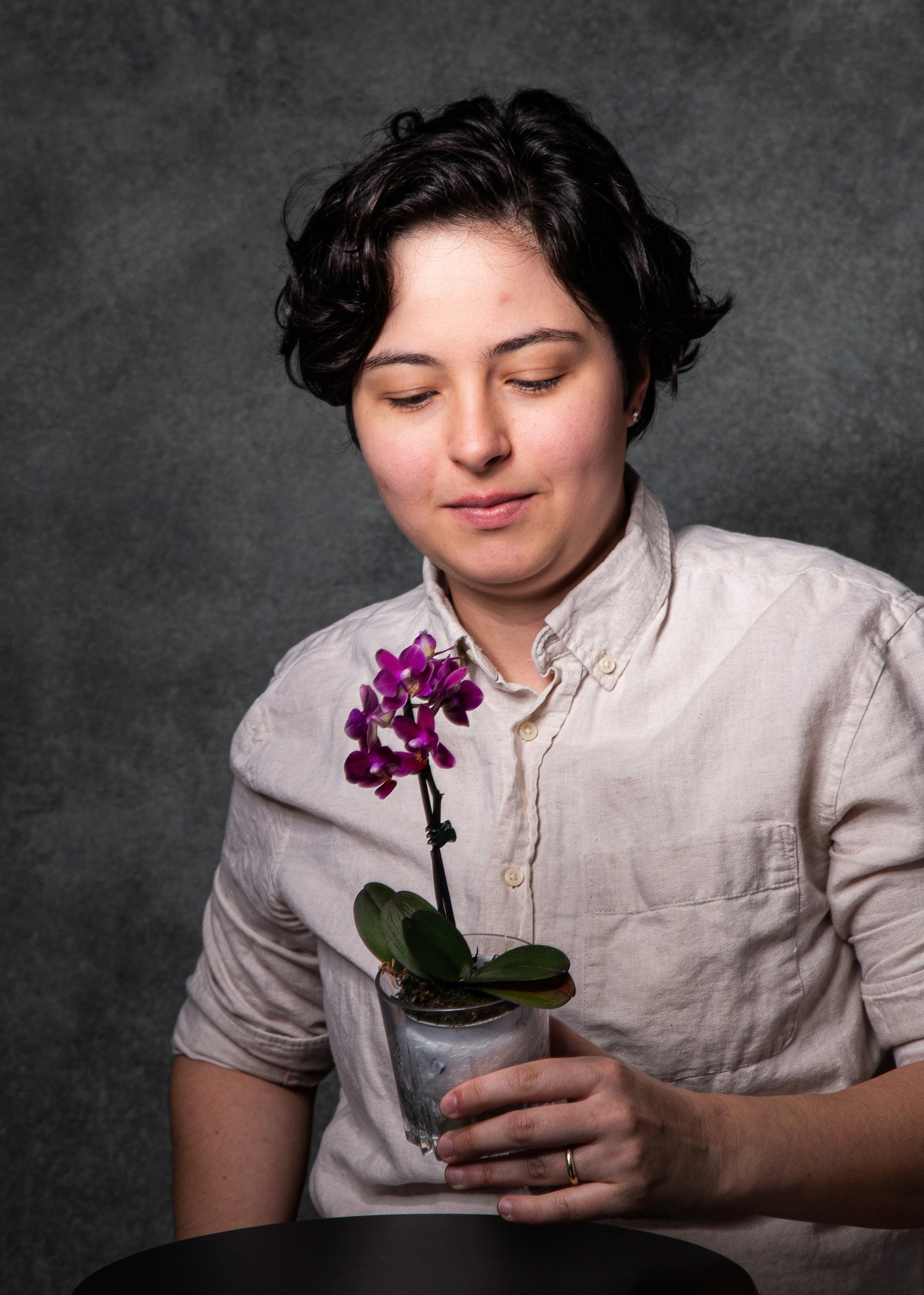 Pessoa de cabelos curtos e escuros, camisa clara, segura pequeno vaso com orquídea roxa e observa a flor, fundo cinza escuro.